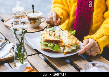 Frau, die im Café im Freien an der City Street frühstückte. Sandwich und Kaffee werden auf dem Tisch serviert. Frau in Strickpullover beim morgendlichen Restaurant Stockfoto
