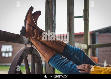 Die Frau entspannt sich auf der Ranch mit ihren Beinen auf einem Holzgeländer. Lederstiefel und Jeans Stockfoto