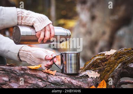 Frau mit Strickhandschuh gießt ein heißes Getränk aus Thermoskanne in den Becher. Erfrischung beim Wandern. Stockfoto