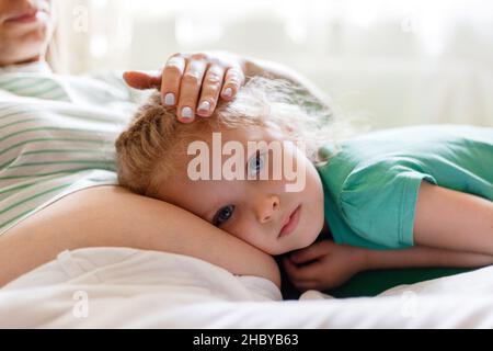 Die Tochter hört dem Herzschlag des Babys zu, wenn sie sich in der Bauchumarmung der Schwangeren umarmt. Porträt der Tochter Kind Kind und schwanger Mutter. Die Zärtlichkeit eines Elternteils und Stockfoto