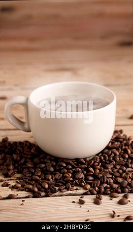 Weiße Tasse Kaffee mit Rauch auf rustikalem Holztisch, mit gerösteten Kaffeebohnen Stockfoto
