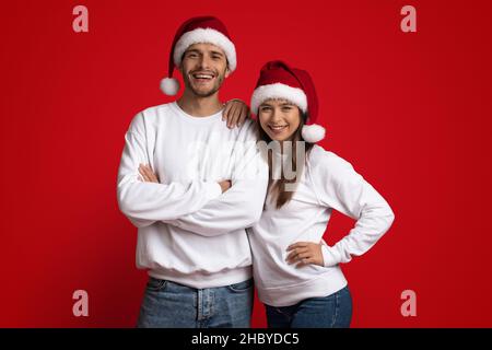 Fröhlicher Junger Mann Und Frau In Santa Hats, Die Auf Rotem Hintergrund Posieren Stockfoto