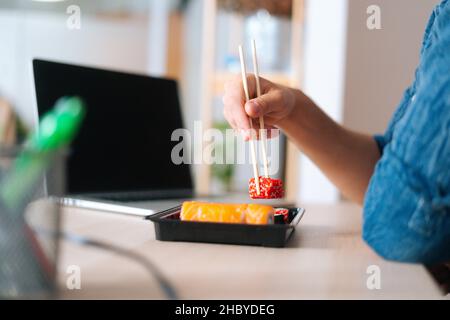 Nahaufnahme von nicht erkennbaren jungen Geschäftsreisenden, die Sushi mit Essstäbchen am Schreibtisch mit Laptop im Heimbüro halten, Stockfoto