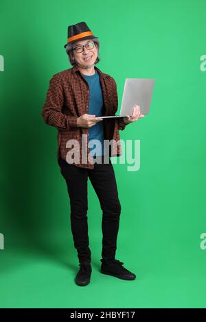 Der ältere asiatische Mann trägt ein orangefarbenes kariertes Hemd und einen schwarzen Hut, der auf dem grünen Hintergrund steht. Stockfoto
