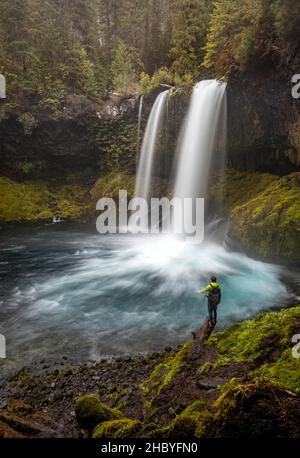 Junger Mann, der auf einem Stein steht, Wasserfall in dichter Vegetation, Koosah Falls, Oregon, USA Stockfoto