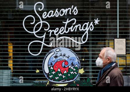 London, Großbritannien. 22. Dezember 2021. Ein Mann mit Gesichtsmask geht in Seven Dials an Weihnachtsdekorationen vor einem geschlossenen Geschäft vorbei. Hospitality-Unternehmen sehen sich in der Zeit, in der sie normalerweise am meisten frequentierten Zeit des Jahres sind, mit reduzierten Einnahmen konfrontiert, da die Kunden aufgrund der Omicron-Variante Buchungen stornieren. Rishi Sunak, Schatzkanzler, hat Pläne für einen £1bn-Fonds angekündigt, um Unternehmen zu helfen, die von dem Anstieg der Covid-Fälle betroffen sind, einschließlich des Freizeit- und Gastgewerbes. Kredit: Stephen Chung / Alamy Live Nachrichten Stockfoto