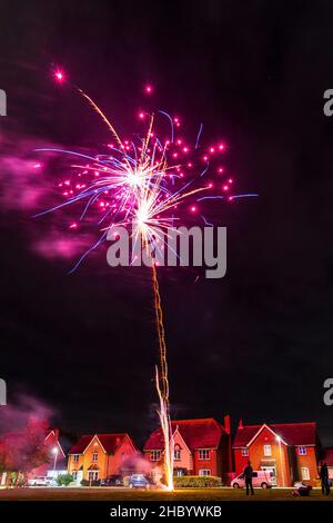 Feuerwerk auf Dorfgrün mit Feuerwerk explodiert auf einer goldenen feurigen Spur in abstraktem, verschwommenem Muster am Himmel über einer Reihe von Häusern. Stockfoto