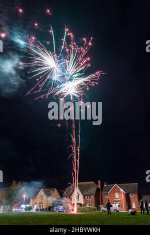 Feuerwerk auf Dorfgrün mit Feuerwerk explodiert auf einer goldenen feurigen Spur in abstraktem, verschwommenem Muster am Himmel über einer Reihe von Häusern. Stockfoto