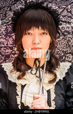 Tokio, Harajuku. Cosplay. Nahaufnahme eines Porträts einer jungen Japanerin in klassisch gotischer Lolita-Kleidung mit Regenschirm, die den Betrachter direkt anschaut. Stockfoto