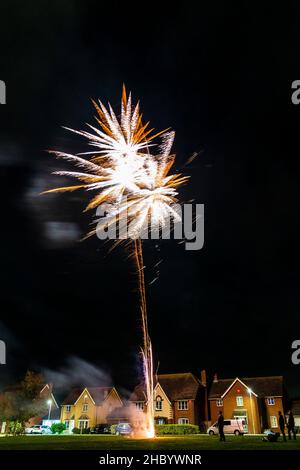 Feuerwerk auf Dorfgrün mit Feuerwerk explodiert auf einer goldenen feurigen Spur in abstraktem, verschwommenem Muster am Himmel über einer Reihe von Häusern. Stockfoto