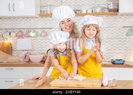 30 Jahre alte kaukasische Mutter hilft ihren süßen kleinen Töchtern beim Rollen von Teig. Stockfoto