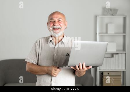 Porträt eines glücklichen leitenden Büromitarbeiters mit einem Laptop. Ein älterer Mann mit grauem Bart hat im Heimbüro einen Job in einem Laptop. Reifer Mann mit Computer Stockfoto