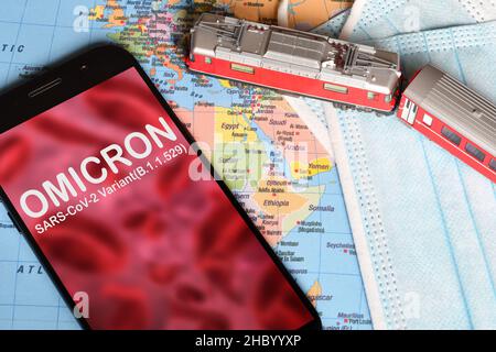Omicron-Variante. Smartphone mit „Omicron“-Text auf abstrakter Unschärfe-Darstellung von roten Viren mit Modelleisenbahn, Schutzmasken und Karten. SARS-CoV-2, B.1.1.52 Stockfoto