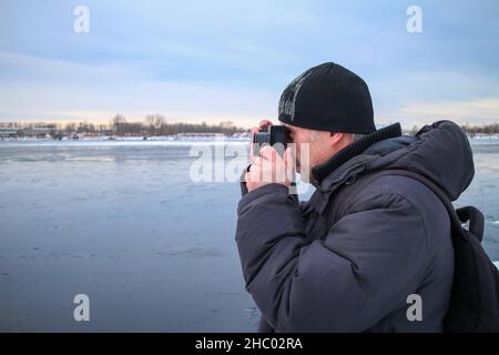 Ein Mann fotografiert im Winter bei kaltem Wetter mit einer Filmkamera. Der Fotograf schaut in den Sucher der Kamera, um den Winter zu fotografieren Stockfoto