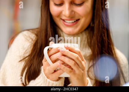 Beschnittenes Foto einer hübschen jungen Dame in warmweißem Pullover, die in der kalten Jahreszeit eine Tasse Tee hält, um sich warm zu halten. Stockfoto