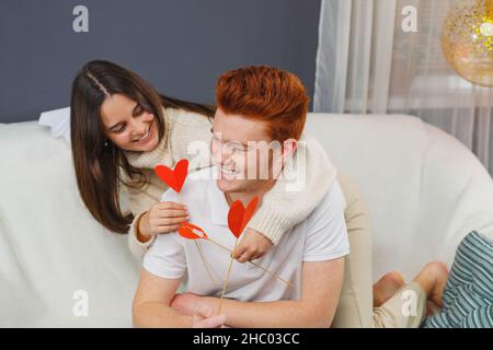 Hübsche junge Dame im warmen Pullover sitzt auf den Knien auf dem Sofa hinter ihrem Freund und flirtet mit herzförmigen Blumen. Stockfoto