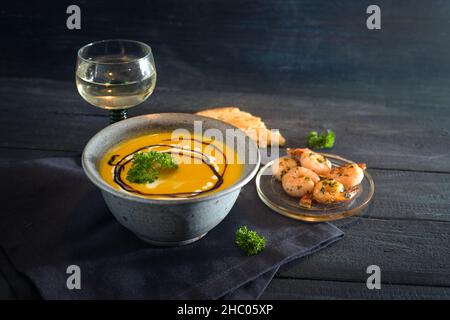 Gemüsesuppe aus rotem Kürbis in einer rustikalen blauen Schüssel mit Garnelen, Petersilie und Weißwein auf einem dunklen Holztisch, Herbstgericht für den Tanksgiv Stockfoto