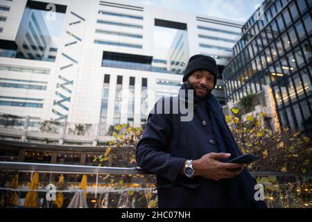 Lächelnder afroamerikanischer Geschäftsmann, elegant in einem schwarzen Mantel gekleidet und trägt an einem Wintertag eine Mütze, hält sein Telefon in der Hand und trägt ein W Stockfoto