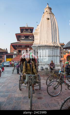 Ein Fahrrad-Rikscha-Fahrer in einem Dhaka-Topi-Hut posiert auf seiner Rikscha auf dem Hanuman Dhoka Durbar Square in Kathmandu, Nepal. Der weiße, sikhara-artige kam D Stockfoto