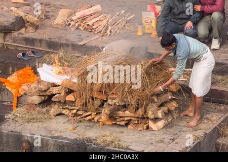 Arbeiter häufen Stroh auf den Körper, während er auf dem Scheiterhaufen des Pashupatinath-Tempelkomplexes in Kathmandu, Nepal, brennt. Stockfoto