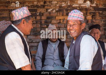 Eine Gruppe nepalesischer Männer in den traditonalen Dhaka-Topi-Kappen ruhen im Schatten und besuchen die mittelalterliche Stadt Bhaktapur in Nepal. Stockfoto