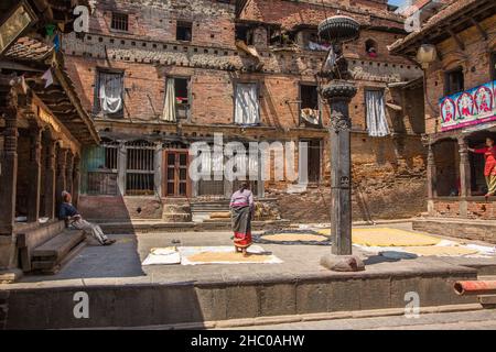 Eine nepalesische Frau geht in der mittelalterlichen Stadt Bhaktapur in Nepal auf ungeschälten Reis, um ihn zu drehen und trocknet in der Sonne. Vorne ist ein stute von Garuda. Stockfoto