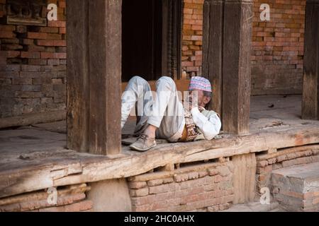 In der mittelalterlichen Stadt Bhaktapur, Nepal, klatscht ein nepalesischer Mann in traditioneller Kleidung und Dhaka-Topi-Mütze im Schatten. Stockfoto