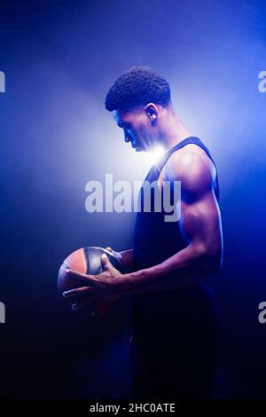 Basketballspieler hält einen Ball vor blauem Nebel Hintergrund. afroamerikanischer Mann, der auf Korb schaut. Stockfoto