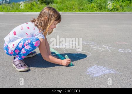 Ein kleines Mädchen zeichnet an einem sonnigen Tag mit Buntstiften auf den Asphalt. Stockfoto