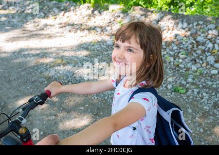 Glückliches Porträt eines kleinen Mädchens mit Fahrrad im Park auf dem Hintergrund einer unbefestigten Straße. Stockfoto