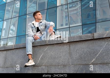 Hübscher junger Mann in stilvoller Kleidung sitzt auf Glasfassade Hintergrund. Junger Mann in der Stadt. Student ruht sich nach dem Studium aus. Stockfoto