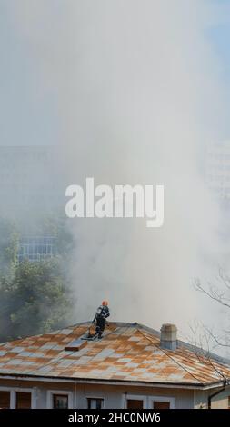 Feuerwehrleute, die auf das Dach des Hauses gehen, um das Feuer aus dem Gebäude in Flammen zu löschen. Feuerwehrmänner der Feuerwehr, deren Ausrüstung versucht, mit Smog und Rauch Flammen aus dem brennenden Haus zu löschen Stockfoto