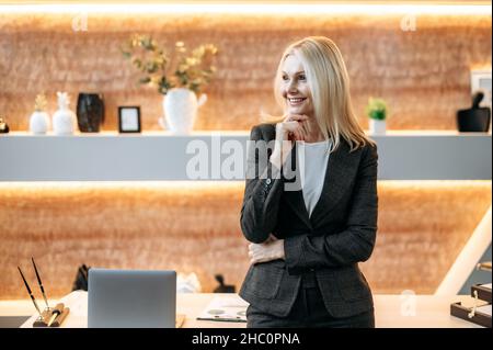 Portrait nachdenklich niedlich glücklich smart erfolgreiche kaukasische reife blonde Geschäftsfrau, ceo oder Top-Manager im modernen Büro in formeller Kleidung zur Seite stehend, Arme gekreuzt, lächelnd Stockfoto