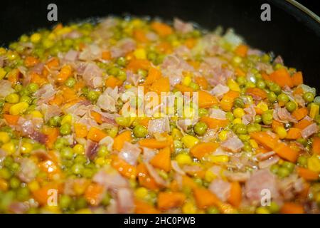 Nahaufnahme eines Gemüserührens Braten Sie mit Karotten, Mais, Erbsen, Zwiebeln und Schinken, in Olivenöl in einer schwarzen Pfanne gebraten, selektiver Fokus Stockfoto