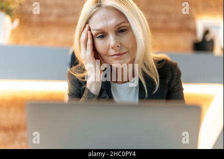 Verschlafen abgemagerte ältere Frau, Geschäftsfrau oder Managerin, in stilvollem Anzug, müde von der Arbeit an Laptops, mit Stress und Kopfschmerzen, massiert ihren Tempel, braucht Ruhe, schaut träge auf den Bildschirm Stockfoto