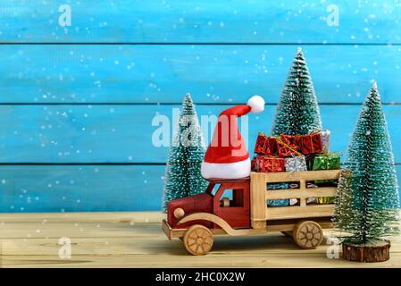 Weihnachtsgeschenke auf einem To Truck mit Weihnachtsmann-Hut neben Tannenbäumen, während es auf einem Holztisch schneit. Stockfoto