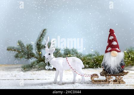 Schlitten mit Weihnachtsgnom und weißen Rentieren neben Tannenzweig, während es auf einem Holztisch schneit. Weihnachts- und Neujahrskonzept. Stockfoto