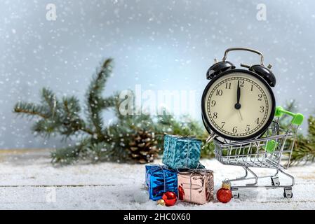 Weihnachtseinkaufskonzept. Wecker in einem Einkaufswagen und Geschenkboxen neben Tannenzweigen, während es auf einem Holztisch schneit. Stockfoto