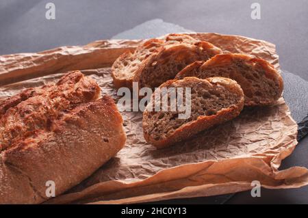 Rustikales Brot, schöne goldene Kruste, geschnittenes Brot auf schwarzem Brett zum Servieren auf Pergamentpapier. Stockfoto