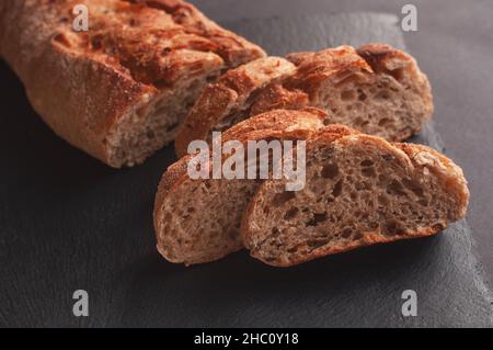 Rustikales Brot, wunderschöne goldene Kruste, Brotscheiben, die zum Servieren auf schwarzem Brett liegen Stockfoto