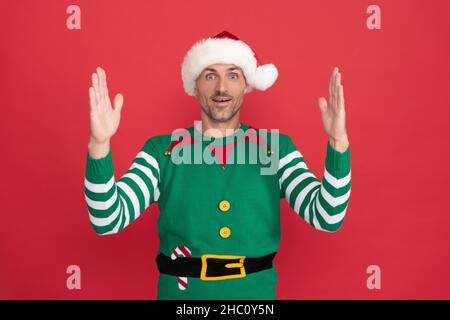 Staunender Mann im Elf Kostüm. weihnachtsmann im weihnachtsmann Hut auf rotem Hintergrund. Frohes neues Jahr. Stockfoto