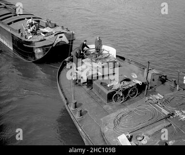 Die seine in Paris führt zu einer Barkacke, von der aus Menschen von einer Brücke herabblicken, 1945. Ein detailliertes Foto, das nur das hintere Ende eines führenden Barges und das vordere Ende des Barges dahinter zeigt. Ein Mann auf dem Frontkahn steuert das überdimensionierte horizontale Steuerruder, während ein zweiter Mann neben ihm steht. Auf der ersten Barge sind ein Fahrrad und eine Weinflasche mit Kork zu sehen. Seillängen säumen das Deck. An der Vorderseite des Schleppkahns zeigt das Bild drei Menschen, die einfach nur mitfahren. Der Fotograf stand auf der Pont de la Concorde, als er dies nahm. Stockfoto