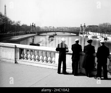 Paris Pont de la Concorde – Männer der US-Marine beobachten im Jahr 1945 die vorbeiführende Barge. Drei Seeleute der Marine und ein Chief Petty Officer stehen und blicken nach Westen über die seine in Richtung Pont Alexandre III und darüber hinaus. Der Schleppkahn ist dabei, unter der Brücke, auf der sie stehen, zu passieren, während er mindestens fünf Lastkähne dahinter zieht. Der Eiffelturm ist im linken Hintergrund und der Palais de Chaillot ist hinten in der Mitte. Die Marinemänner (sowie der Fotograf Clarence Inman) sind jeweils im Pariser Außenbüro des Field Photo Branch, O.S.S., stationiert Stockfoto