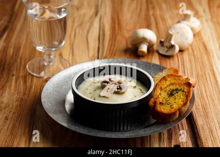 Hausgemachte, frisch zubereitete, cremige Pilzsuppe mit knusprigem Knoblauchbrot Baguette, auf schwarzer Keramikschüssel oder Ramequin über dem rustikalen Tisch servieren. Stockfoto