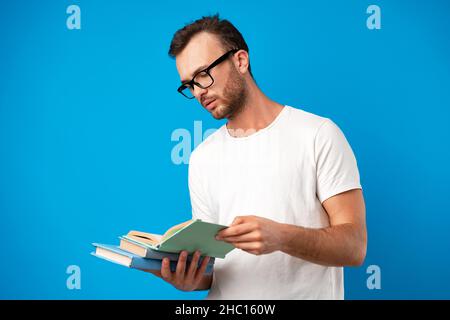 Junger Mann mit Brille, der vor blauem Hintergrund steht und ein Buch liest Stockfoto