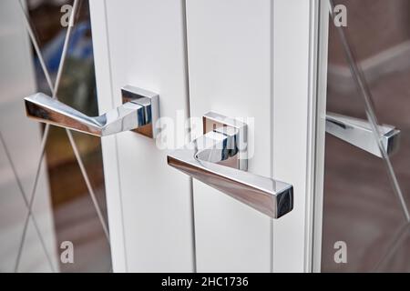 Glänzende silberne Griffe an geschlossener moderner Zimmertür mit gemustertem Spiegelglas in elegant renovierter Wohnung extreme close view Stockfoto