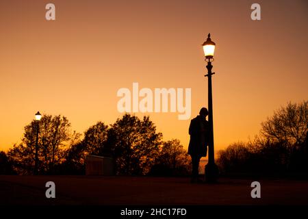 Eine Einzelfigur eines Mannes, der sich an einem Laternenpfosten lehnte, und gegen einen Abendhimmel auf Primrose Hill, London, geschildet wurde Stockfoto