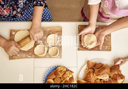 Ältere Frau mit erwachsenen Tochter knetet und rollt rohen Teig und kocht leckere Kuchen am weißen Tisch in der Küche oben aus der Nähe Stockfoto