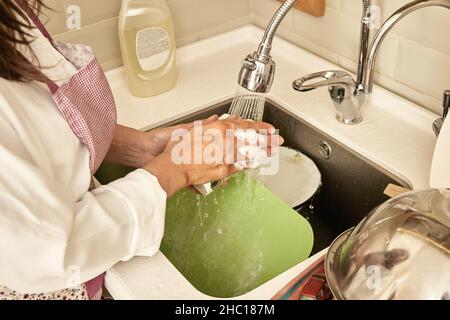 Frau in Schürze und schäumende Hände wäscht schmutziges Geschirr mit fließendem Wasser im modernen Waschbecken in der hellen Küche aus nächster Nähe Stockfoto