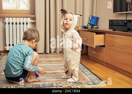 Niedliches Baby trägt einen warmen Hirsch-Body mit Hörnern und ein älterer Bruder spielt mit einer Konstruktion, die auf dem Boden im geräumigen Wohnzimmer aufgestellt ist Stockfoto